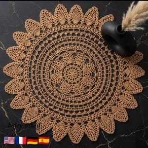 May include: A circular, tan crochet doily with a decorative scalloped border. The detailed openwork pattern includes concentric circles and a central floral design. A black vase with dried grass is in the background. The text "Enjoy 50% off" is at the bottom.