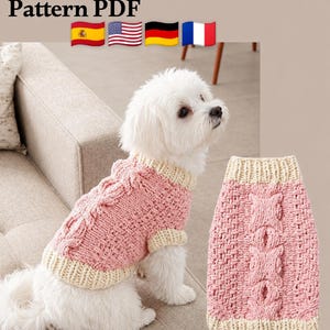 May include: A Pattern PDF for a dog sweater featuring a pink cable knit design with cream-colored ribbed trim at the neck, armholes, and hem. The sweater is shown worn by a small white dog and also displayed separately as a flat garment.