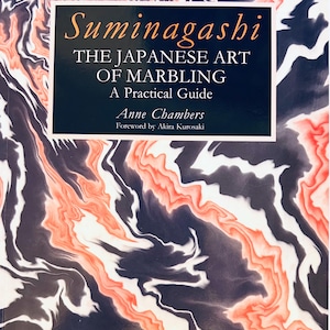 May include: A book titled "Suminagashi: The Japanese Art of Marbling: A Practical Guide" by Anne Chambers. The cover features a marbled design in shades of black, white, and orange. The title is in orange, and the author's name is in white.