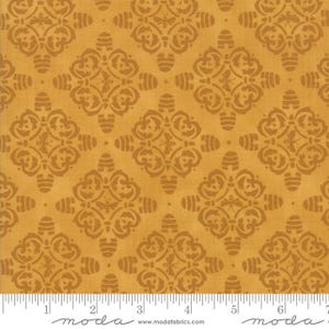 May include: Mustard-colored fabric with a repeating pattern of ornate, stylized floral and geometric shapes in a darker shade of brown. The fabric has a textured appearance and is suitable for quilting or other craft projects. The image includes a ruler along the bottom.
