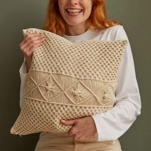 Op de afbeelding: Een beige macramé kussen met een geometrisch ontwerp. Het kussen heeft een geweven textuur aan de boven- en onderkant, met een decoratieve band van ruit- en bloemvormen in het midden. Het kussen wordt vastgehouden door een persoon die een wit shirt met lange mouwen en een beige broek draagt.