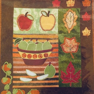 May include: A square fabric wall hanging featuring an autumn theme with appliqued motifs. The design includes a red apple, a yellow apple, a bowl of green pears, sliced fruit, orange pumpkins on a vine, and various colorful fall leaves.