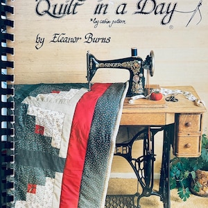 May include: A vintage Singer sewing machine sits on a wooden table next to a partially visible quilt. The quilt features a log cabin pattern with red, gray, and white blocks. The book cover reads "Make a Quilt in a Day" by Eleanor Burns.