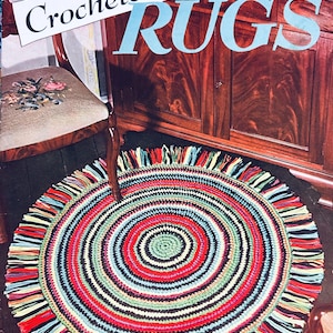May include: A vintage magazine cover titled "Crocheted Rugs" features a colorful, round rug with a spiral design. The rug has a fringe and is made of red, green, blue, black, and white yarn. The magazine cover is in the background.
