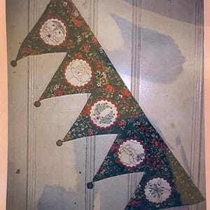 May include: A Christmas banner featuring four triangular pennants. Each pennant is green with a floral pattern and a circular embroidered design with festive motifs. Brown ball ornaments hang from the tips of the pennants.