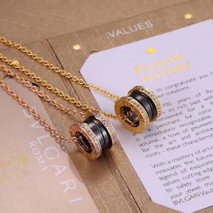May include: Two necklaces with pendant charms. One is gold-toned, the other rose gold. Each pendant features a black band with a ring of clear crystals. The necklaces are displayed on a card with the text "BVLGARI HISTORY".