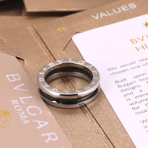 May include: A silver ring with the brand name "BVLGARI" engraved around the outer edge. The ring has a black band in the center and is displayed on a light brown surface with cards.