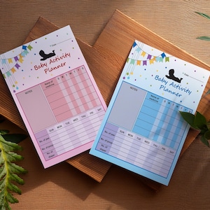 May include: Two baby activity planners, one pink and one blue, with a banner design and the text "Baby Activity Planner." Each planner has sections for notes, medicine, and diaper changes. The planners are on a wooden surface.