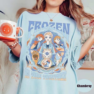 May include: Light blue t-shirt featuring the text "FROZEN IN THIS TOGETHER" with a graphic of characters from the Disney movie Frozen. The shirt has a crew neck and short sleeves. A white and orange mug with a sun design is held in the left hand.