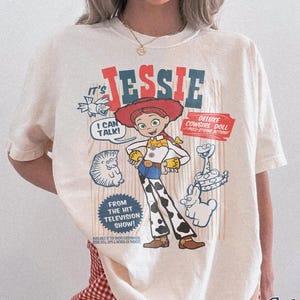 Peut inclure: T-shirt ivoire avec un imprim&eacute; de Jessie de Toy Story. Le motif comprend le texte "It's Jessie", "I Can Talk!" et "Deluxe Cowgirl Doll". Le t-shirt est &eacute;tiquet&eacute; "Comfort Colors" et a une coupe d&eacute;contract&eacute;e.