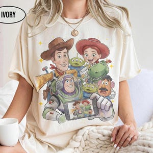 May include: Ivory-colored t-shirt featuring a colorful graphic of characters from a popular animated movie. The design includes Woody, Buzz Lightyear, Jessie, and other characters. The shirt has short sleeves and a crew neck.