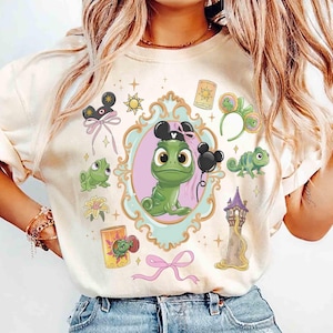 May include: A cream-coloured t-shirt featuring a central graphic of a green chameleon inside an ornate light blue frame. The design includes a tall tower, a yellow lantern, a flower, a pink bow, and various small icons like mouse ears and stars.
