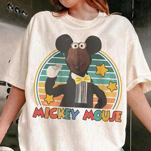 May include: Ivory-colored t-shirt featuring a cartoon mouse in a tuxedo, bow tie, and gloves, set against a retro striped background with stars. The words "MICKEY MOUSE" are printed in colorful letters below the image.