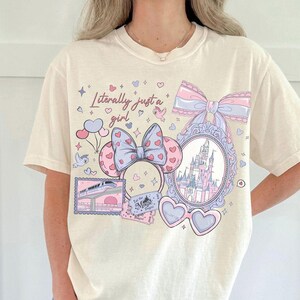 May include: Cream-colored t-shirt with a whimsical graphic. The design includes a castle, bows, heart-shaped glasses, and the phrase "Literally just a girl." The graphic is in pastel pinks, blues, and purples, creating a soft, dreamy aesthetic. The shirt has a relaxed fit.