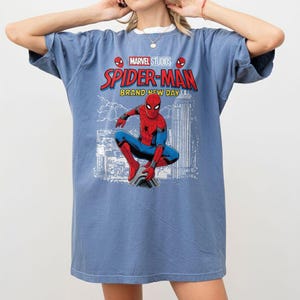 Pode incluir: Camiseta azul com um gr&aacute;fico do Homem-Aranha da Marvel Studios. O design inclui o texto "Spider-Man Brand New Day" e um cen&aacute;rio urbano. A camisa tem um ajuste casual e grande.
