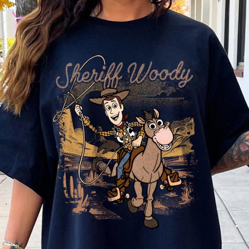 May include: Navy blue t-shirt featuring a graphic of Sheriff Woody from Toy Story riding a horse. The text "Sheriff Woody" is arched across the top in a glittery gold font. The design is set against a desert landscape.