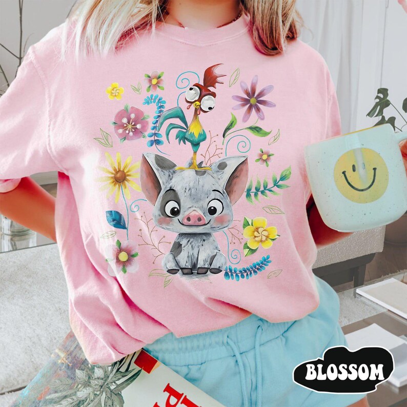 May include: Pale pink t-shirt with a cartoon pig, rooster, and floral design. The shirt has a crew neck and short sleeves. The word "BLOSSOM" is printed in a black oval at the bottom right.