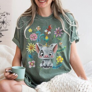 May include: Green t-shirt with a cartoon pig wearing a crown, alongside a rooster and flowers. The design is printed on a soft tee, suitable for everyday wear. The shirt has a relaxed fit and is a comfortable choice.