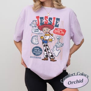Peut inclure: T-shirt lilas avec un imprim&eacute; de Jessie, personnage de Toy Story. Le motif comprend le texte "Jessie", "I can talk!" et "Deluxe Cowgirl Doll". Le t-shirt est &eacute;tiquet&eacute; "Comfort Colors Orchid".