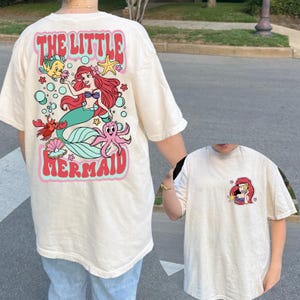 May include: Cream-colored t-shirt featuring a colorful graphic of Ariel from The Little Mermaid. The back of the shirt displays the words "THE LITTLE MERMAID" in a retro font, along with cartoon characters. A smaller version of Ariel is on the front.
