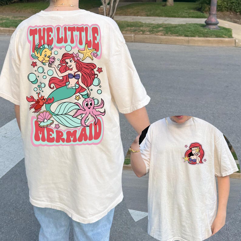 May include: Cream-colored t-shirt featuring a colorful graphic of Ariel from The Little Mermaid. The back of the shirt displays the words "THE LITTLE MERMAID" in a retro font, along with cartoon characters. A smaller version of Ariel is on the front.