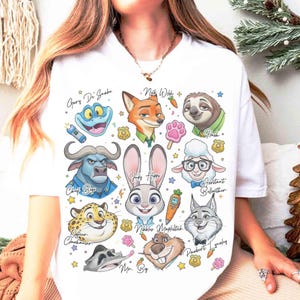 K&ouml;nnte beinhalten: Wei&szlig;es T-Shirt mit farbenfrohen Cartoon-Illustrationen von Charakteren aus Zootopia, darunter Nick Wilde, Judy Hopps und Flash. Das Design enth&auml;lt Charakternamen und verschiedene thematische Symbole. Ein lustiges, auff&auml;lliges Grafik-T-Shirt.