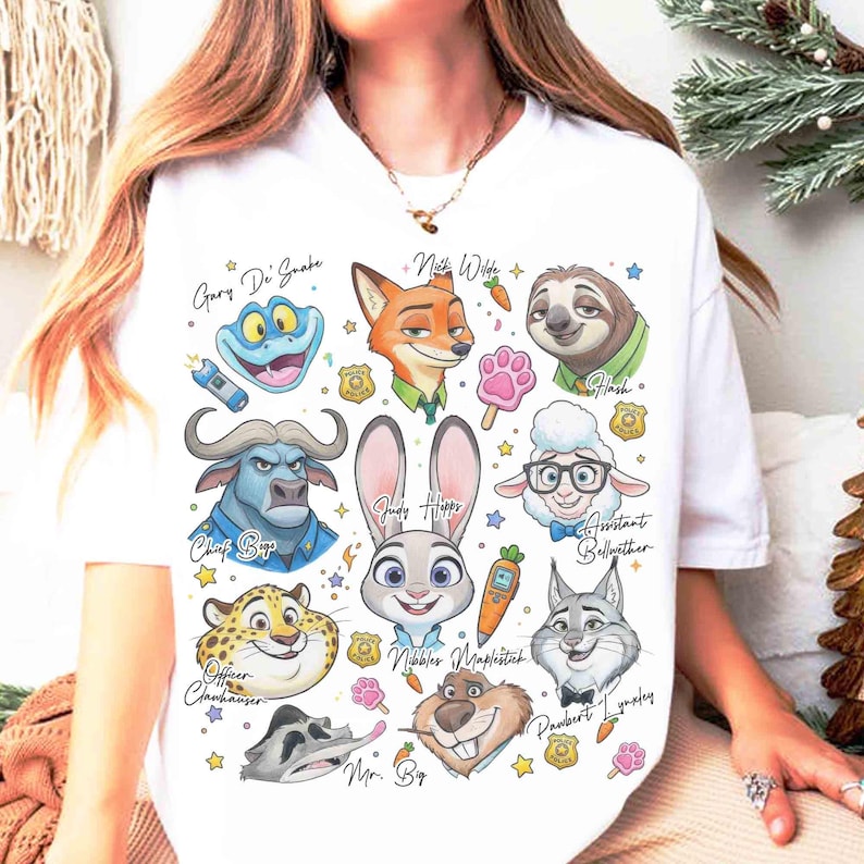 K&ouml;nnte beinhalten: Wei&szlig;es T-Shirt mit farbenfrohen Cartoon-Illustrationen von Charakteren aus Zootopia, darunter Nick Wilde, Judy Hopps und Flash. Das Design enth&auml;lt Charakternamen und verschiedene thematische Symbole. Ein lustiges, auff&auml;lliges Grafik-T-Shirt.