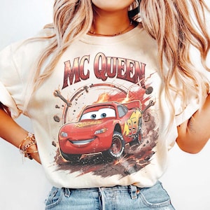 May include: Cream-colored t-shirt featuring a graphic of Lightning McQueen from the movie Cars. The design includes the text "MC QUEEN" above the car and the number "95" on the side. The car is red with yellow accents and is depicted in a dynamic, action-oriented pose.