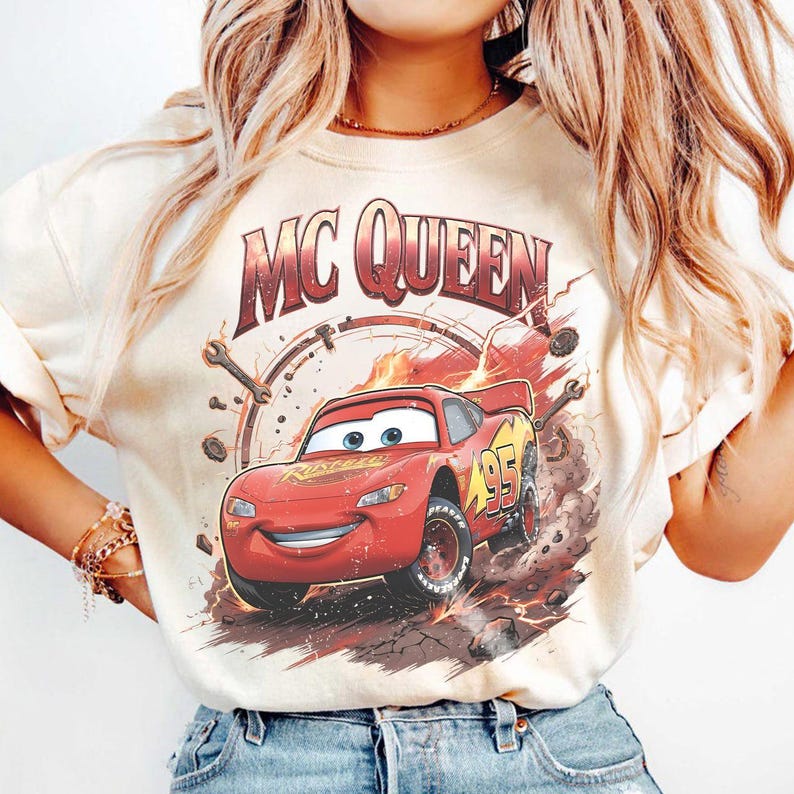 May include: Cream-colored t-shirt featuring a graphic of Lightning McQueen from the movie Cars. The design includes the text "MC QUEEN" above the car and the number "95" on the side. The car is red with yellow accents and is depicted in a dynamic, action-oriented pose.