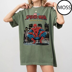 May include: Olive green t-shirt featuring a graphic of Spider-Man in a crouching pose against a cityscape backdrop. The text "MARVEL STUDIOS SPIDER-MAN BRAND NEW DAY" is printed above the image in red and blue.