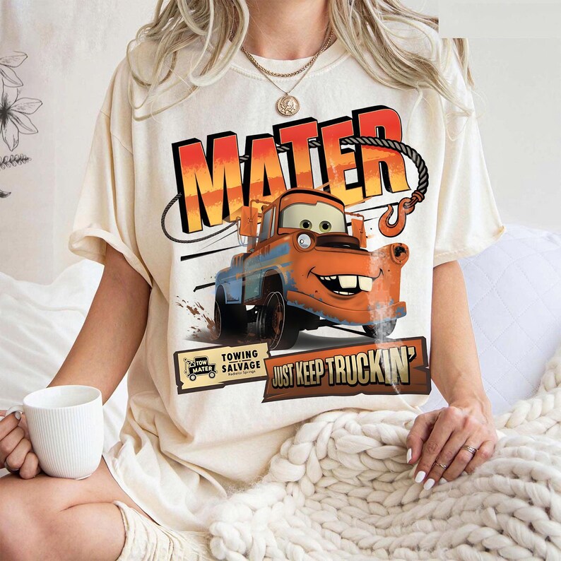 Puede incluir: Camiseta color crema con un gr&aacute;fico de Mate de la pel&iacute;cula Cars. El dise&ntilde;o incluye la palabra "MATER" en naranja y rojo, y la frase "JUST KEEP TRUCKIN'". La camiseta tambi&eacute;n tiene un peque&ntilde;o gr&aacute;fico que dice "Towing Salvage".