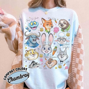K&ouml;nnte beinhalten: Hellblaues Sweatshirt mit Zeichentrickfiguren aus Zootopia, darunter Judy Hopps, Nick Wilde und Flash. Das Shirt tr&auml;gt den Schriftzug "COMFORT COLORS Chambray" in Schreibschrift.