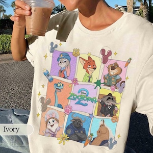 May include: Ivory-colored t-shirt featuring a colorful graphic of characters from the Disney movie Zootopia. The design includes Judy Hopps, Nick Wilde, and other characters in a grid pattern with the movie's logo. The shirt is made of a soft material.