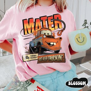 Puede incluir: Camiseta rosa claro con un dise&ntilde;o de Mate de la pel&iacute;cula Cars. El dise&ntilde;o incluye el nombre "MATER" en naranja y rojo, y la frase "JUST KEEP TRUCKIN'". Tambi&eacute;n se ve una taza azul claro con una carita sonriente amarilla.