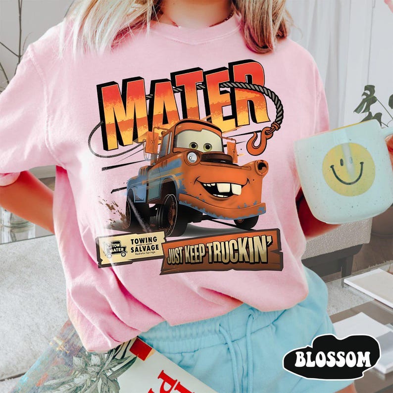 Puede incluir: Camiseta rosa claro con un dise&ntilde;o de Mate de la pel&iacute;cula Cars. El dise&ntilde;o incluye el nombre "MATER" en naranja y rojo, y la frase "JUST KEEP TRUCKIN'". Tambi&eacute;n se ve una taza azul claro con una carita sonriente amarilla.
