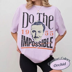 May include: Orchid-colored t-shirt with the text "Do The Impossible" and a portrait. The year "1955" is printed above the word "Impossible". The shirt has a Comfort Colors label.