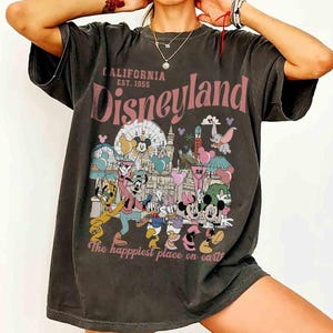May include: Dark grey t-shirt with a Disneyland graphic. The design features the words "California Disneyland Est. 1955" in pink, along with cartoon characters and park imagery. The phrase "The happiest place on earth" is also included.