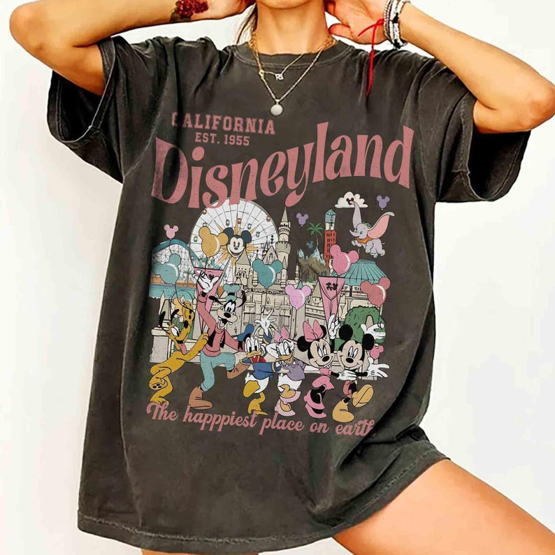 May include: Dark grey t-shirt with a Disneyland graphic. The design features the words "California Disneyland Est. 1955" in pink, along with cartoon characters and park imagery. The phrase "The happiest place on earth" is also included.