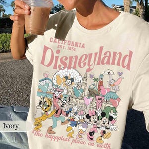 May include: Ivory t-shirt with a Disneyland graphic. The design features the text "CALIFORNIA EST. 1955 Disneyland" in pink, alongside cartoon characters and park imagery. The phrase "The happiest place on earth" is also included.