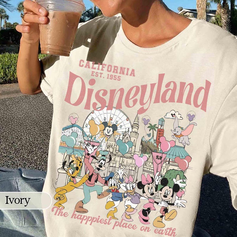 May include: Ivory t-shirt with a Disneyland graphic. The design features the text "CALIFORNIA EST. 1955 Disneyland" in pink, alongside cartoon characters and park imagery. The phrase "The happiest place on earth" is also included.