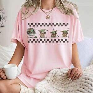 May include: A light pink t-shirt featuring a graphic of a character from a popular science fiction franchise. The graphic shows the character in four different poses, with a black and white checkered pattern above and below the graphic.