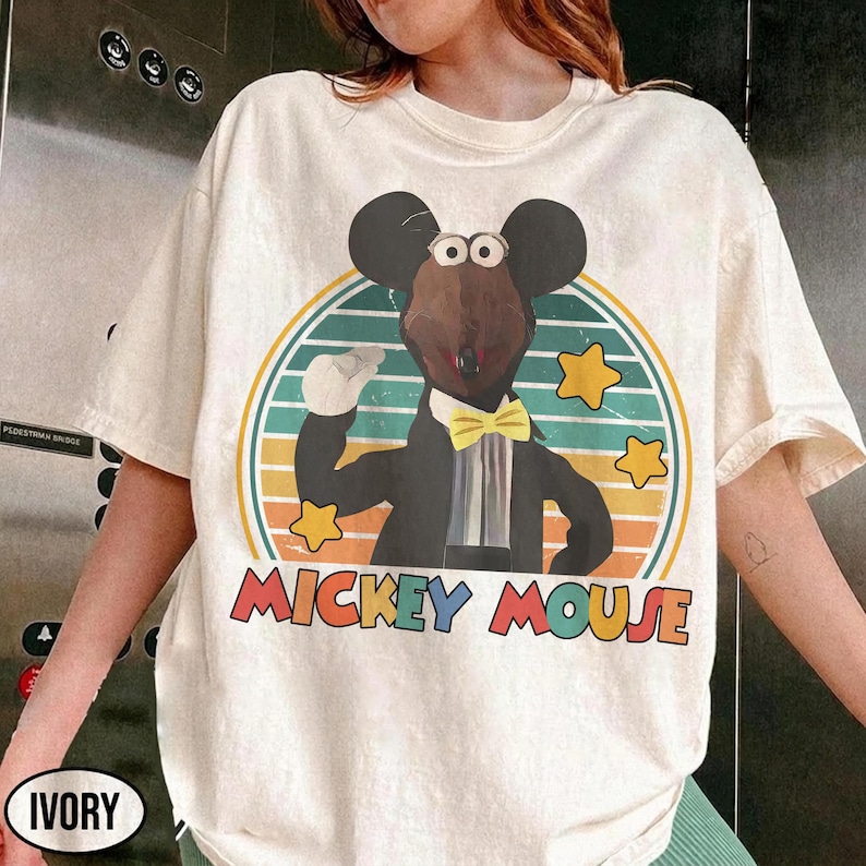 May include: Ivory-colored t-shirt featuring a cartoon mouse in a tuxedo, bow tie, and gloves, set against a retro striped background with stars. The words "MICKEY MOUSE" are printed in colorful letters below the image.