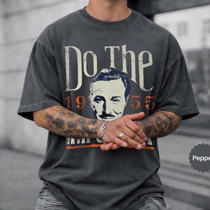 May include: Pepper-colored t-shirt with the text "Do The" above a vintage portrait and the year "1955". The shirt has a distressed look, with an orange stripe at the bottom. The shirt is a short-sleeved, crew neck style.