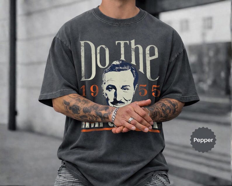 May include: Pepper-colored t-shirt with the text "Do The" above a vintage portrait and the year "1955". The shirt has a distressed look, with an orange stripe at the bottom. The shirt is a short-sleeved, crew neck style.