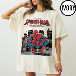 May include: Ivory-colored t-shirt featuring a Marvel Studios Spider-Man graphic. The design includes the words "Spider-Man Brand New Day" above an illustration of Spider-Man in a crouching pose against a city skyline. The shirt has short sleeves.