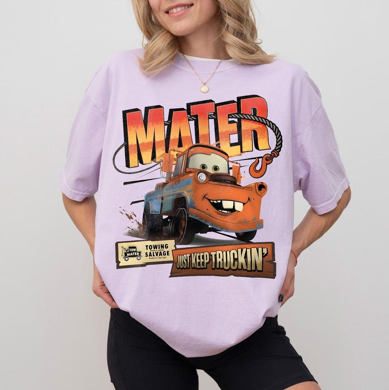 Puede incluir: Una camiseta de color lila claro con un dise&ntilde;o de Mate de la pel&iacute;cula Cars. El dise&ntilde;o incluye el nombre "MATER" en letras grandes y la frase "JUST KEEP TRUCKIN'". La camiseta tambi&eacute;n tiene un peque&ntilde;o logotipo que dice "TOW MATER SALVAGE".