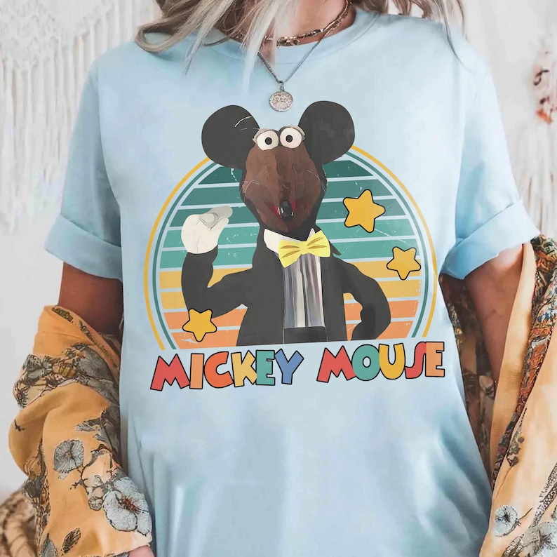 May include: Light blue t-shirt featuring a cartoon mouse character in a tuxedo with a yellow bow tie. The design includes a retro sunset background and the words "MICKEY MOUSE" in colorful letters. The shirt has rolled-up sleeves.