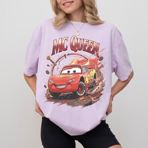 May include: A light purple t-shirt featuring a graphic of Lightning McQueen from the movie Cars. The design includes the text "MC QUEEN" above the car and the number 95 on the side. The car is red with yellow accents.