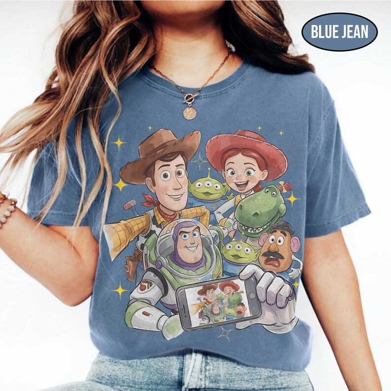 May include: Blue jean colored t-shirt featuring a graphic of Toy Story characters, including Woody, Buzz Lightyear, and Jessie. The characters are arranged in a group, with some holding a phone displaying a photo. The text "BLUE JEAN" is in a blue oval.