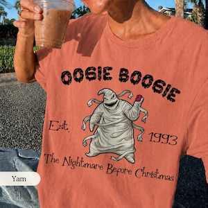 Puede incluir: Una camiseta de color terracota con un gr&aacute;fico del personaje Oogie Boogie. El texto dice OOGIE BOOGIE, Est. 1993 y The Nightmare Before Christmas. El personaje est&aacute; representado con una forma de saco gris claro y una amplia sonrisa dentada.